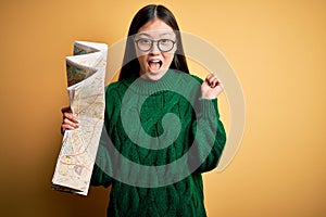 Young asian turist woman looking at city tourist map on a trip over yellow background screaming proud and celebrating victory and