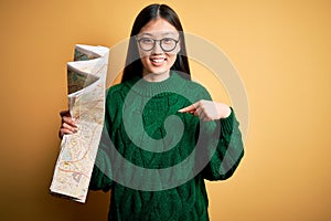 Young asian turist woman looking at city tourist map on a trip over yellow background with surprise face pointing finger to