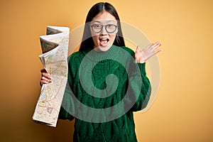 Young asian turist woman looking at city tourist map on a trip over yellow background very happy and excited, winner expression