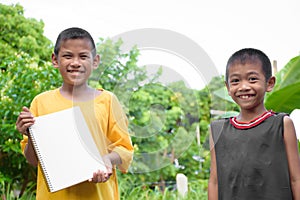 Young Asian two boys yellow and black t-shirt smiling and holding a blank white notebook on a blurred forest and tree.