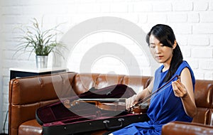 Young asian violinist in blue dress  takes care of the instrument after  practice session in living room. Rubbing rosin on the