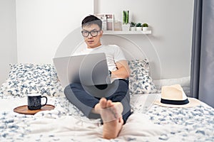 Young Asian wearing eyes glasses and working at home. A man sitting on bed and working with laptop computer on bed. Work from home
