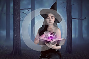 Young asian wizard woman holding spell book in the forest with f