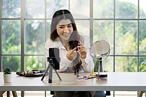 Young asian woman is applying lipstick, recording a makeup tutorial with a smartphone and mirror