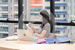 Young asian woman in casual with face mask and note of schedule on the calendar. New normal office with desk, laptop, documents