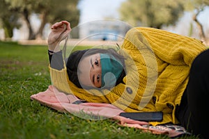 Young Asian woman in city park during covid19 - happy and pretty Korean girl in face mask lying relaxed on green grass during