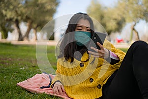 Young Asian woman in city park during covid19 - happy and pretty Korean girl in face mask using internet with mobile phone relaxed