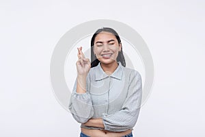 A young asian woman closes her eye wishing for good luck. Crossing her fingers hoping for the best. Isolated on a white background