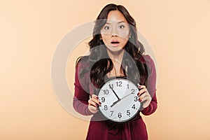 Young asian woman with curly long hair holding clock showing nearly 8 being late or missing something over peach background