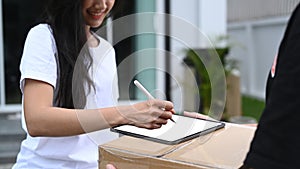 Young Asian woman customer signing on digital tablet and receiving package from delivery man.