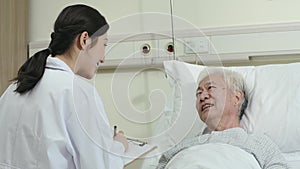 Young asian woman doctor talking to senior male patient in hospital ward