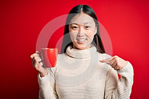 Young asian woman drinking a cup of hot coffee over isolated red background with surprise face pointing finger to himself