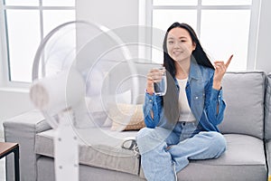 Young asian woman drinking glass of water enjoying air from fan smiling happy pointing with hand and finger to the side