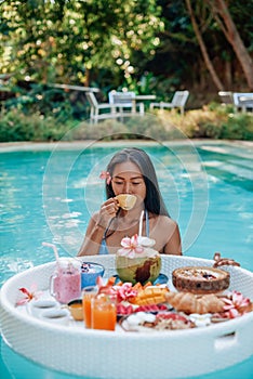 Young asian woman drinks fruit juice swimming in pool with floating table