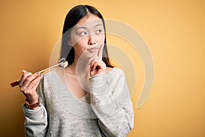 Young asian woman eating japanese food, holding salmon and rice maki sushi using chopsticks serious face thinking about question,