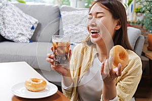 young asian woman enjoy eating sweet donut and drink cola soft drink in living room at home in summer season