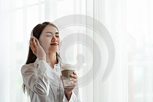 Young Asian woman enjoy listening to music with headphones and holding a hot cup of coffee, happy and relaxing time