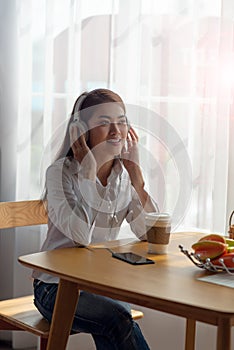 Young Asian woman enjoy listening to music with headphones and a hot cup of coffee on the table, happy and relaxing time