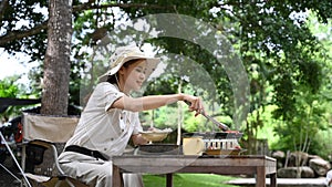 Young asian woman enjoy making barbecue during camping near the river bank. Outdoor activity or vacation concept