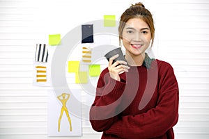 Young asian woman fashion designer holding coffee cup and smiling while standing over sketch paper and fabric on studio office