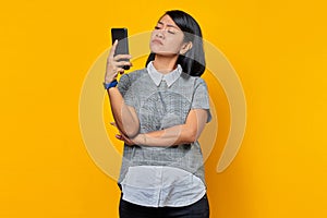 Young Asian woman feeling annoyed and angry while looking at message on smartphone on yellow background