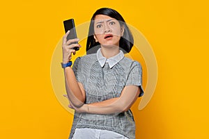 Young Asian woman feeling annoyed and angry while looking at message on smartphone on yellow background