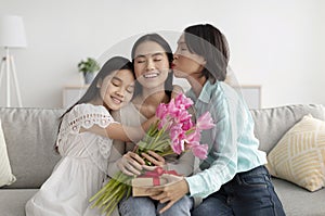 Young Asian woman with flowers and gift box being kissed and hugged by her mature mom and cute little daughter at home