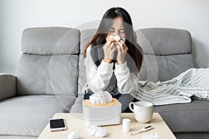 Young Asian woman get sick with changing seasons sitting on couch. Girl feel cold and has a high fever blowing nose