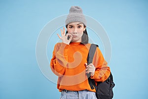 Young asian woman, girl zips her mouth, tells secret, stands over blue studio background