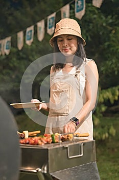 Young asian woman grilling barbeque skewers meat on bbq grill in backyard on picnic time