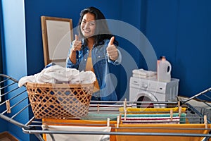 Young asian woman hanging clothes at clothesline approving doing positive gesture with hand, thumbs up smiling and happy for