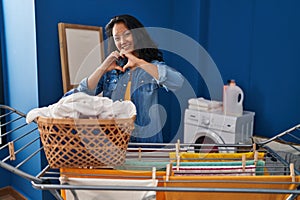 Young asian woman hanging clothes at clothesline smiling in love doing heart symbol shape with hands
