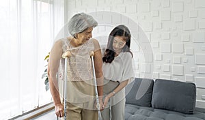 Young asian woman help her grandma walk by using axilla crutches. Senior gray hair woman exercise and practice walking at home