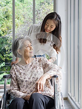 Young asian woman help pushing a wheelchair for grandma in front of corridor with glass wall