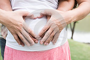 Young asian woman and her husband together caressing her pregnant belly holding their hands in a heart shape on her baby bump. Pr