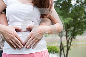 Young asian woman and her husband together caressing her pregnant belly holding their hands in a heart shape on her baby bump. Pr