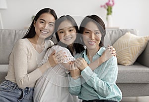 Young Asian woman with her mature mother and adorable little daughter hugging and looking at camera at home
