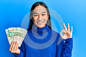 Young asian woman holding 50 hong kong dollars banknotes doing ok sign with fingers, smiling friendly gesturing excellent symbol