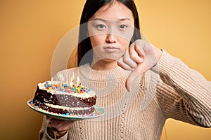 Young asian woman holding birthday cake with candles burning over yellow isolated background with angry face, negative sign