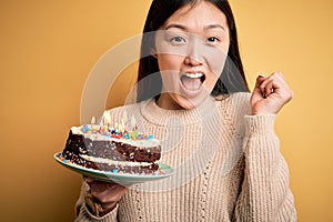 Young asian woman holding birthday cake with candles burning over yellow isolated background screaming proud and celebrating