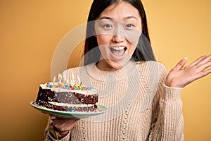 Young asian woman holding birthday cake with candles burning over yellow isolated background very happy and excited, winner