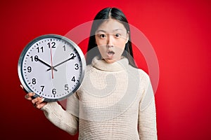 Young asian woman holding countdown big clock over red isolated background scared in shock with a surprise face, afraid and