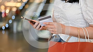 Young Asian woman holding her credit card, using smartphone, walking in the shopping mall