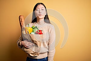 Young asian woman holding paper bag of fresh healthy groceries over yellow  background looking at the camera blowing a