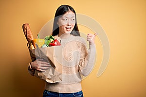 Young asian woman holding paper bag of fresh healthy groceries over yellow  background smiling with happy face looking and