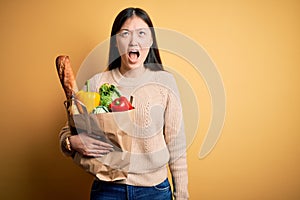 Young asian woman holding paper bag of fresh healthy groceries over yellow isolated background angry and mad screaming frustrated