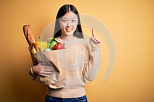 Young asian woman holding paper bag of fresh healthy groceries over yellow isolated background with a big smile on face, pointing