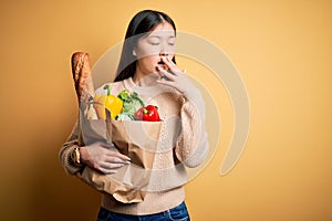 Young asian woman holding paper bag of fresh healthy groceries over yellow isolated background bored yawning tired covering mouth