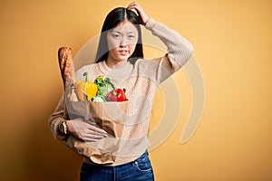 Young asian woman holding paper bag of fresh healthy groceries over yellow isolated background confuse and wondering about