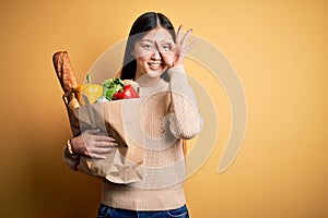 Young asian woman holding paper bag of fresh healthy groceries over yellow isolated background doing ok gesture with hand smiling,